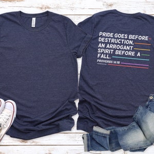 May include: A navy blue t-shirt with a white graphic print that reads "PRIDE GOES BEFORE DESTRUCTION, AN ARROGANT SPIRIT BEFORE A FALL. PROVERBS 16:18". The graphic is in a rainbow color scheme.
