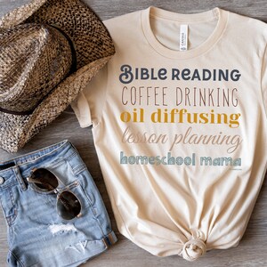 May include: A beige t-shirt with the text "Bible Reading, Coffee Drinking, Oil Diffusing, Lesson Planning, Homeschool Mama" in colourful lettering. The shirt is laid out on a wooden surface with a straw cowboy hat, denim shorts, and brown ankle boots.