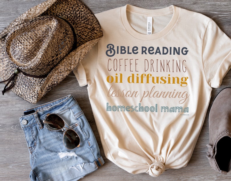 May include: A beige t-shirt with the text "Bible Reading, Coffee Drinking, Oil Diffusing, Lesson Planning, Homeschool Mama" in colourful lettering. The shirt is laid out on a wooden surface with a straw cowboy hat, denim shorts, and brown ankle boots.