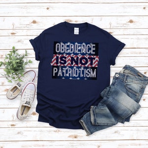 May include: A navy blue t-shirt displaying the message "OBEDIENCE IS NOT PATRIOTISM" in white lettering. The text is set against a patriotic design of red and white stripes and blue stars. The shirt is shown with blue jeans and white trainers.