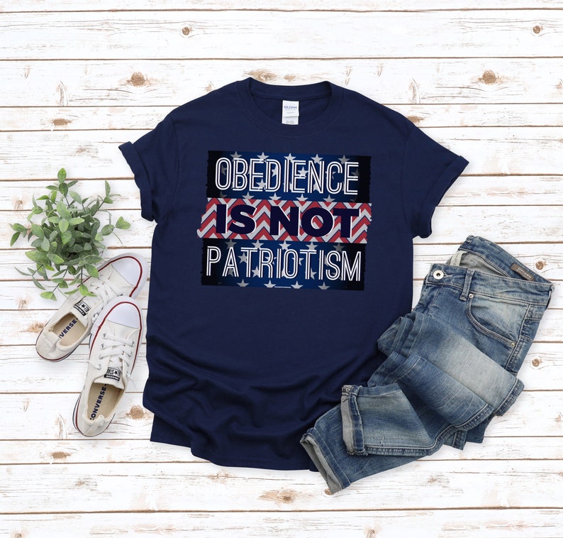 May include: A navy blue t-shirt displaying the message "OBEDIENCE IS NOT PATRIOTISM" in white lettering. The text is set against a patriotic design of red and white stripes and blue stars. The shirt is shown with blue jeans and white trainers.