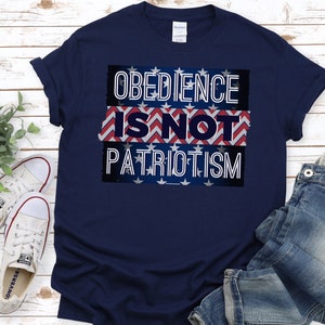 May include: A navy blue t-shirt featuring the white text "OBEDIENCE IS NOT PATRIOTISM" over a red, white, and blue design. The shirt is shown on a light wood surface, with white trainers and blue jeans visible.