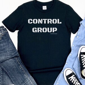 May include: Black t-shirt with white text that reads "CONTROL GROUP". The shirt is laid flat on a white surface with blue jeans, a light blue button-down shirt, and black sneakers.