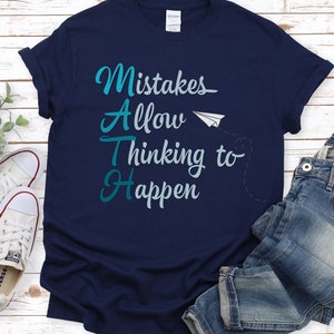Pode incluir: Camiseta azul marinho com um gráfico de avião de papel branco e o texto "Mistakes Allow Thinking to Happen" em letras azul turquesa.