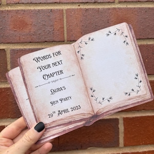 May include: A small, open book-shaped card with a vintage, weathered paper appearance. The left page features the text: Words for your next chapter, Laura's Hen Party, 29th April 2023. The right page has a delicate, black floral vine border.