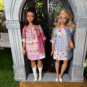 May include: Two dolls standing side by side. The doll on the left has dark hair and wears a pink and white floral patchwork dress with white knee-high boots. The doll on the right has blonde hair and wears a blue and white floral patchwork dress.