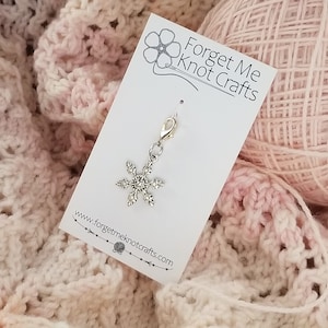 Puede incluir: Marcapuntos de punto de copo de nieve plateado con cierre de langosta. El marcador está en una tarjeta blanca con el texto "Forget Me Knot Crafts" y una dirección de sitio web.