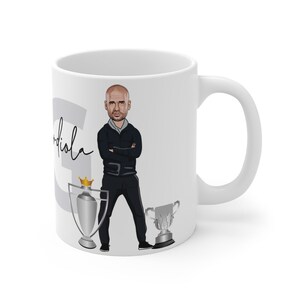 May include: White ceramic coffee mug with a cartoon illustration of a man with a black jacket and black pants. The man is standing with his arms crossed. The mug also has a silver trophy with a gold crown on top and a silver trophy with a gold base. The text "Guardiola" is written on the mug.