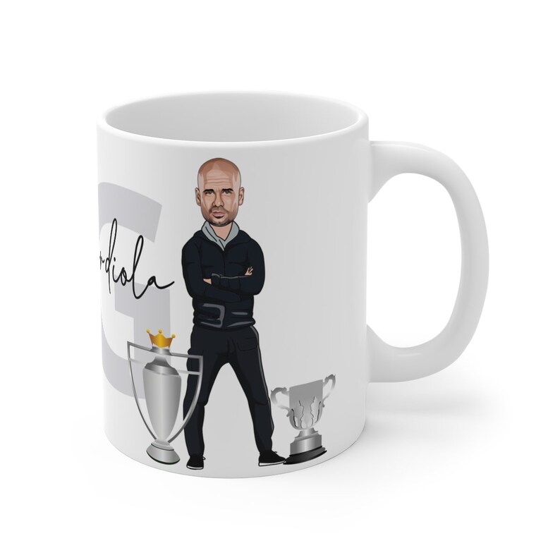 May include: White ceramic coffee mug with a cartoon illustration of a man with a black jacket and black pants. The man is standing with his arms crossed. The mug also has a silver trophy with a gold crown on top and a silver trophy with a gold base. The text "Guardiola" is written on the mug.