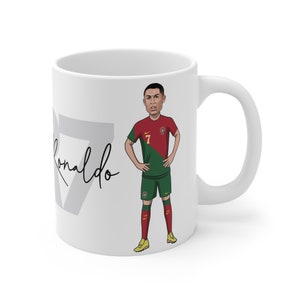 May include: White ceramic mug with a cartoon illustration of a soccer player wearing a red and green jersey with the number 7. The player's name is Cristiano Ronaldo and his signature is written in black on the mug.