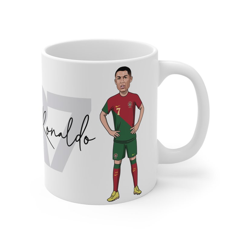 May include: White ceramic mug with a cartoon illustration of a soccer player wearing a red and green jersey with the number 7. The player's name is Cristiano Ronaldo and his signature is written in black on the mug.