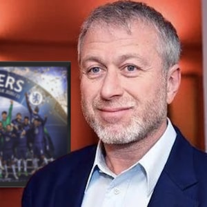 May include: A man in a blue suit smiles, with a framed image of the Chelsea football team celebrating their UEFA Champions League victory. The image on the frame reads "WINNERS UEFA CHAMPIONS LEAGUE 20/21".