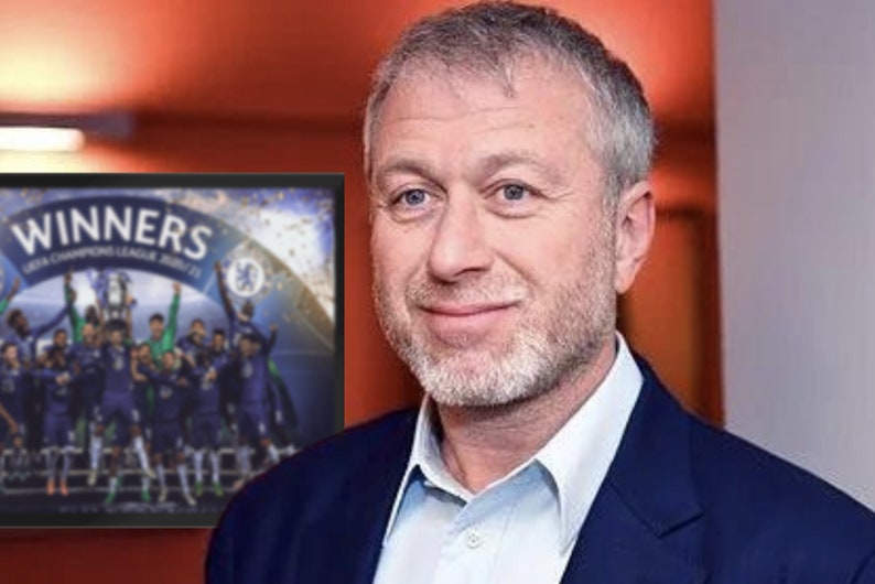 May include: A man in a blue suit smiles, with a framed image of the Chelsea football team celebrating their UEFA Champions League victory. The image on the frame reads "WINNERS UEFA CHAMPIONS LEAGUE 20/21".