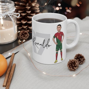May include: White ceramic mug with a cartoon illustration of Cristiano Ronaldo wearing a Portugal soccer jersey. The mug has the text "Ronaldo" printed on the side.