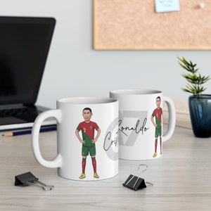 May include: Two white ceramic mugs with a cartoon image of Cristiano Ronaldo in a soccer uniform. The mug on the left has the number 7 and Ronaldo's signature. The mug on the right has the number 7 and the text "Ronaldo".