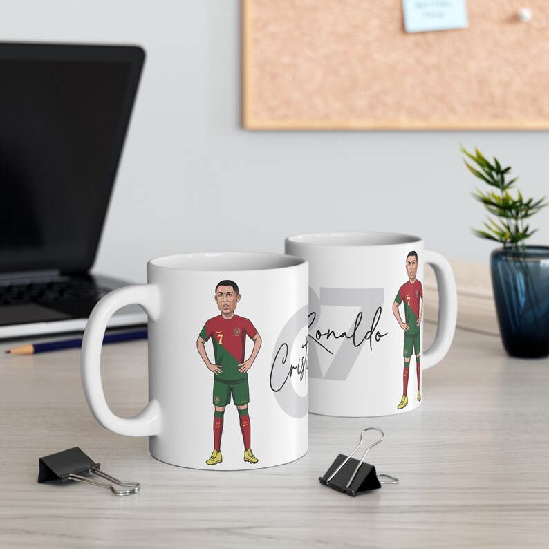 May include: Two white ceramic mugs with a cartoon image of Cristiano Ronaldo in a soccer uniform. The mug on the left has the number 7 and Ronaldo's signature. The mug on the right has the number 7 and the text "Ronaldo".