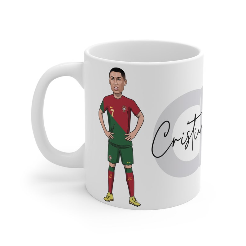 May include: White ceramic mug with a cartoon illustration of Cristiano Ronaldo wearing a Portugal soccer jersey. The mug has a gray circle with the text "Cristiano" in black cursive.