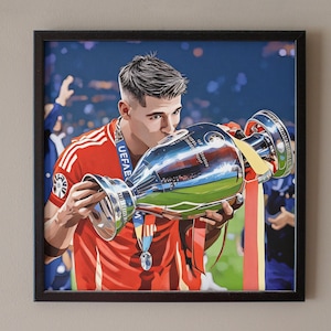 May include: Framed print of a soccer player kissing a trophy. The player wears a red jersey and a UEFA medal. The trophy is silver with a gold interior, reflecting a green field. The background is a blurred stadium.