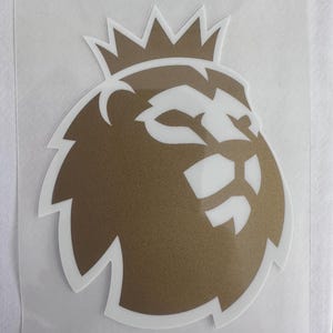 Puede incluir: Una pegatina dorada y blanca con el logotipo de la Premier League. El logotipo es una cabeza de león coronada. La cabeza del león es dorada y los detalles son blancos. La pegatina está sobre un soporte transparente.