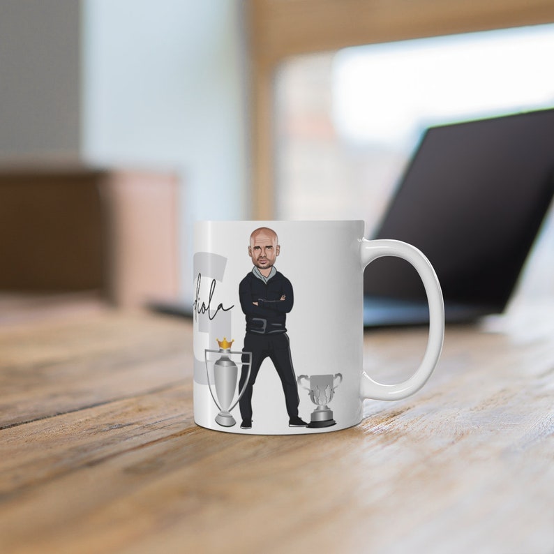 May include: White ceramic mug with a cartoon illustration of a man in a black suit and tie, standing with his arms crossed. The mug has two silver trophies with gold accents on either side of the man. The text "Pep" is written on the mug.