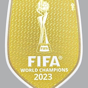 May include: A gold-coloured FIFA World Champions 2023 patch with a white outline. The patch features a trophy with a football on top and the text "FIFA" in white.