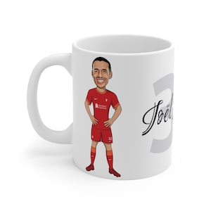 Könnte beinhalten: Weiße Keramiktasse mit einer Cartoon-Illustration eines Fußballspielers, der ein rotes Trikot mit dem Text "Standard Chartered" und der Nummer 32 trägt. Der Name des Spielers "Joël" ist in Kursivschrift auf der Tasse geschrieben.