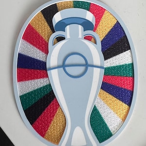 Puede incluir: Un trofeo blanco y azul con un fondo a rayas de colores dorado, rojo, verde, negro y blanco. El trofeo tiene forma de jarrón con dos asas.