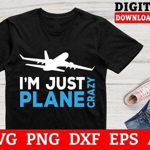 May include: Black t-shirt with the text "I'M JUST PLANE CRAZY" and a white airplane graphic. The words "PLANE" are in blue. The image also includes a pair of pink sneakers and denim shorts.