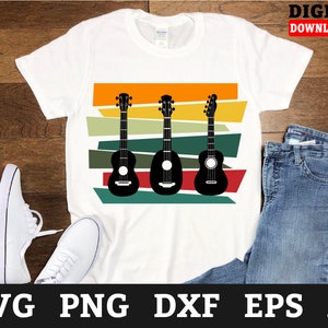 Könnte beinhalten: Ein weißes T-Shirt mit einem Design aus drei schwarzen Ukulelen auf einem bunten gestreiften Hintergrund. Die Ukulelen sind in einer Reihe angeordnet, wobei die Hälse nach rechts zeigen. Der Hintergrund besteht aus horizontalen Streifen in Orange-, Gelb-, Grün- und Brauntönen.