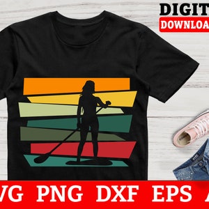 May include: A black t-shirt featuring a silhouette of a person paddleboarding against a sunset backdrop. The design incorporates a retro colour scheme with orange, yellow, green, and red stripes.