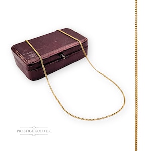 Op de afbeelding: Een goudkleurige ketting over een bordeauxrode sieradendoos. De ketting heeft een delicaat, geschakeld ontwerp. De rechthoekige sieradendoos heeft een sluiting. De woorden "PRESTIGE GOLD UK" staan onderaan de afbeelding.