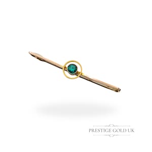 Könnte beinhalten: Eine goldene Bar-Brosche mit einem grünen Edelstein, der in einem runden Rahmen gefasst ist. Die Brosche befindet sich auf einem weißen Hintergrund.