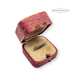 Pode incluir: Caixa de anel bordô antiga e aberta com exterior texturizado. O interior é forrado com um material de veludo bege. A caixa é retangular com tampa articulada e um pequeno fecho. O texto "PRESTIGE GOLD UK" é visível.
