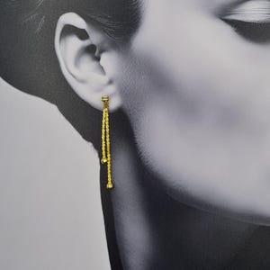May include: A gold-tone drop earring featuring two delicate, textured chains of varying lengths suspended from a small stud post. Each chain terminates in a tiny, smooth gold-tone bead, creating a layered, dangling effect that hangs vertically.