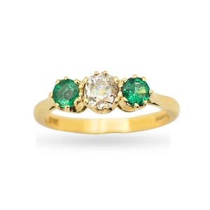 Pode incluir: Um anel de ouro amarelo com uma pedra preciosa branca redonda central, ladeada por duas pedras verdes redondas. As pedras são fixadas por garras numa banda de ouro lisa e polida que se estreita ligeiramente em direção à parte posterior.