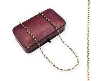 May include: A gold-colored chain necklace draped over a burgundy jewelry box. The box is rectangular with a clasp and a textured surface. The chain is composed of small, linked circles.
