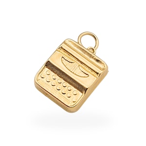 Op de afbeelding: Een goudkleurige bedel in de vorm van een vintage typemachine. De bedel heeft een gedetailleerd toetsenbord met ronde toetsen en een centraal designelement. Er zit een kleine lus aan de bovenkant om aan een ketting of armband te bevestigen. De bedel is gemaakt van glanzend metaal.