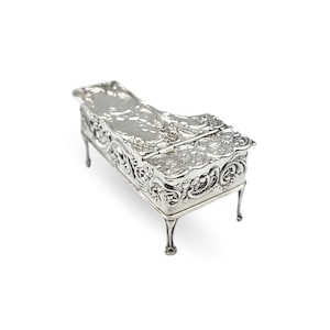 May include: A miniature silver-coloured piano-shaped box, featuring detailed floral and scrollwork. The box has a hinged lid and elegant, curved legs. The design is reminiscent of antique jewellery boxes or decorative items.