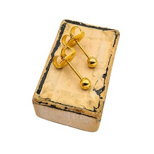 May include: A pair of gold-colored stud earrings with a ball at the end of a slender post. The earrings are displayed on a small, aged, rectangular box with a worn, tan surface and black trim. The background is white.