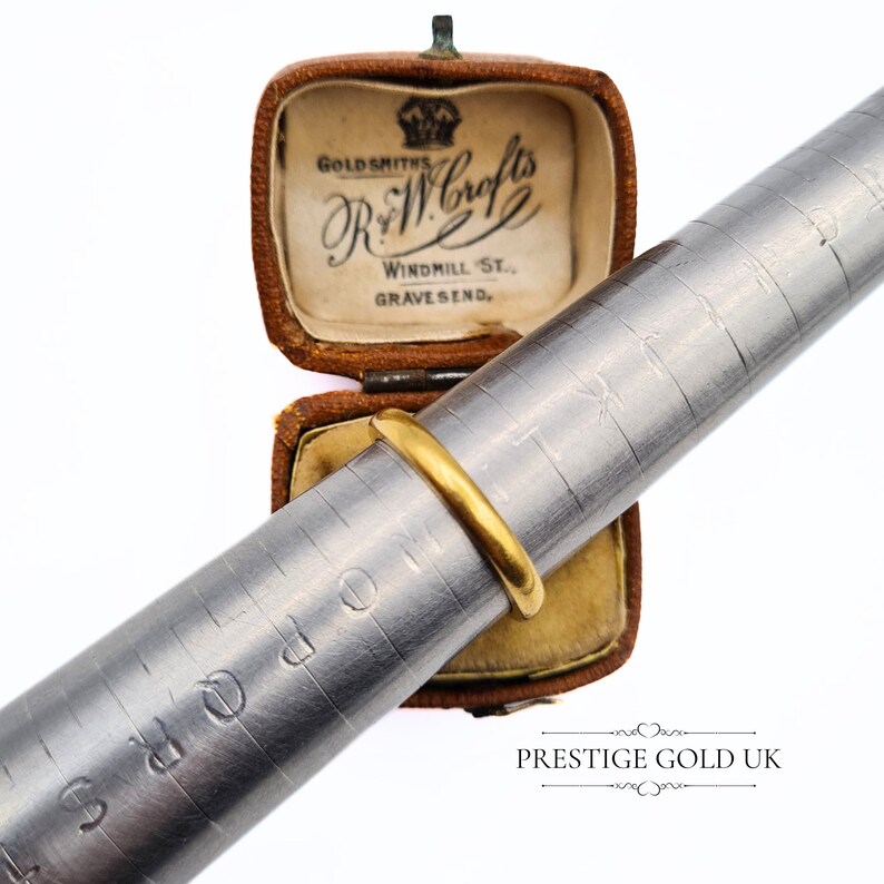 May include: A gold wedding band on a ring sizer. The ring sizer is marked with sizes. The ring is in a small brown box with the inscription "GOLDSMITHS R & W Crofts WINDMILL ST., GRAVESEND."