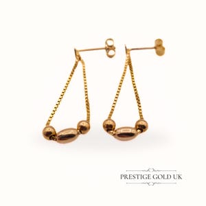 May include: A pair of gold drop earrings with a chain design and three round beads. The earrings have a post back closure.