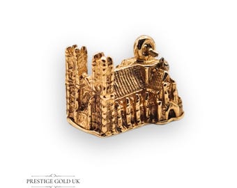 May include: A gold-colored miniature of a cathedral, featuring intricate architectural details. The charm has a detailed roof, arched windows, and a small figure on top. The base of the charm is curved. The text "PRESTIGE GOLD UK" is at the bottom.