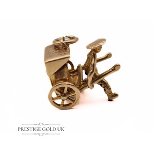 May include: A gold-colored charm depicting a person pulling a rickshaw. The charm is detailed and features a person in a traditional hat and a rickshaw with a single wheel.