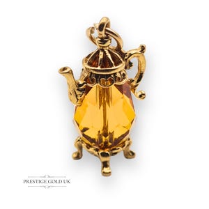 May include: A gold-colored teapot charm with a faceted amber-colored crystal body. The charm has intricate details, including a handle, spout, and a decorative lid. The teapot sits on a small, ornate stand. The text "PRESTIGE GOLD UK" is at the bottom.