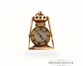 May include: A gold-toned miniature lantern pendant with a clock face. The clock face has black and gold markings and hands. The lantern has a decorative top and side supports. The text "PRESTIGE GOLD UK" is at the bottom.
