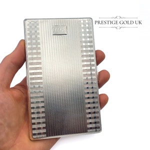 May include: A silver rectangular case with a textured surface and a small rectangular emblem with the initials "D.B.J." The case has a geometric pattern and is held in a hand. The text "PRESTIGE GOLD UK" is in the upper right corner.