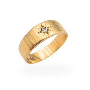 May include: A gold ring with a starburst design and a small, clear gemstone at the center. The ring has a brushed finish and a rounded band. The starburst is slightly raised, adding dimension to the design. The ring is set against a white background.