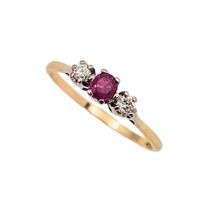 May include: A gold ring featuring an oval ruby flanked by two diamonds. The ring has a classic design with a gold band and white metal settings. The gemstones are set in a prong setting. The ring is a piece of jewellery.