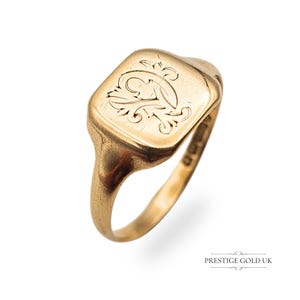 May include: A gold signet ring with a rectangular face. The face features an engraved floral design. The ring is a warm gold color and has a smooth, polished finish. The ring is shown against a white background.