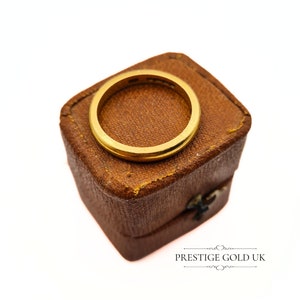 May include: A gold wedding band in a brown velvet ring box. The box is closed and the ring is visible on top.